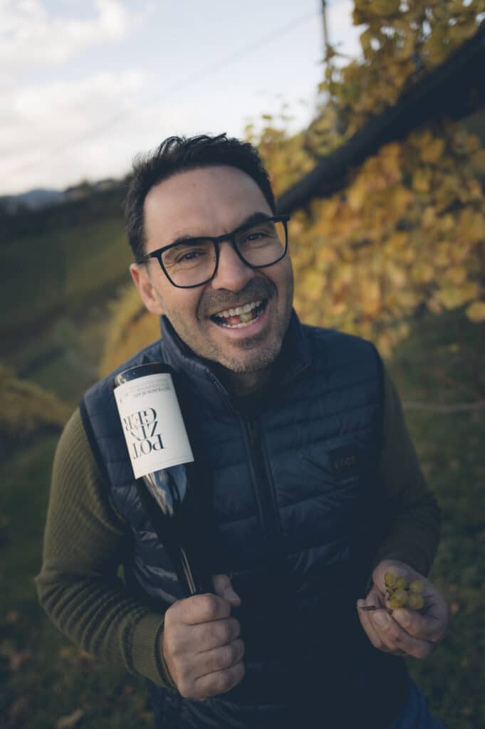 Ein lächelnder Mann mit Brille und dunkler Weste steht in einem Weinberg, in der Hand eine Flasche Wein und eine kleine Traube, im Hintergrund herbstlich gefärbte Weinblätter.