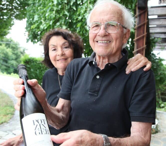 Ein älteres Paar lächelt im Freien, wobei der Mann eine Flasche Wein mit der Aufschrift "Potzinger" in der Hand hält. Beide tragen schwarze Hemden und sitzen an einem Tisch mit Grünzeug und einer Holzkonstruktion im Hintergrund.