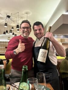 Zwei Männer lächeln in einem Restaurant; einer zeigt den Daumen nach oben, während der andere eine große Weinflasche mit der Aufschrift "Pot Anger" hält. Esstisch und Gläser im Vordergrund.